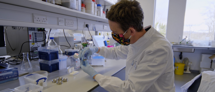 Man wearing face mask sat at a lab bench, wearing a lab coat and holding tools.