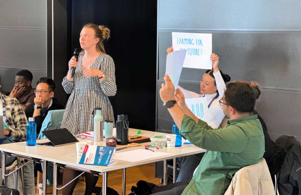 A student stood at a table talking into a microphone with two students holding up signs - one is visible and says Farming for the future