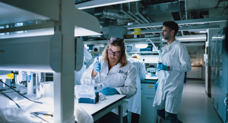 Prof Caroline Gauchotte-Lindsay wearing a lab coat and holding a pipette and test tube in a lab, while three colleagues are working around her, also wearing lab coats