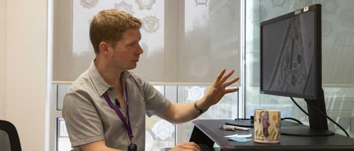 A person standing at a height-adjustable desk gestures towards a computer monitor displaying a grayscale scientific image.