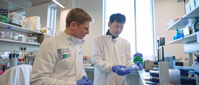 Two researchers wearing white lab coats and blue gloves work together at a laboratory bench. One holds a lab container while the other looks on. The workspace is filled with scientific equipment, supplies, and shelving, with natural light coming through tall windows.