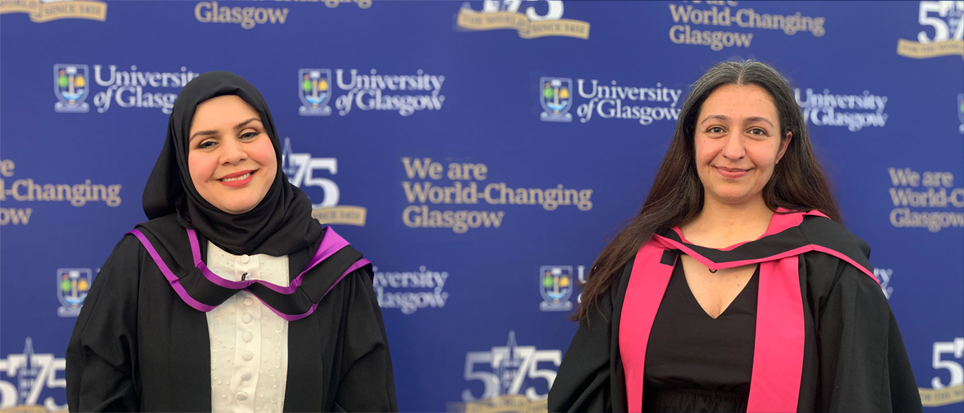 Dr Aksu and Hosay posing for a photo with graduation robes on in front of a university branded board