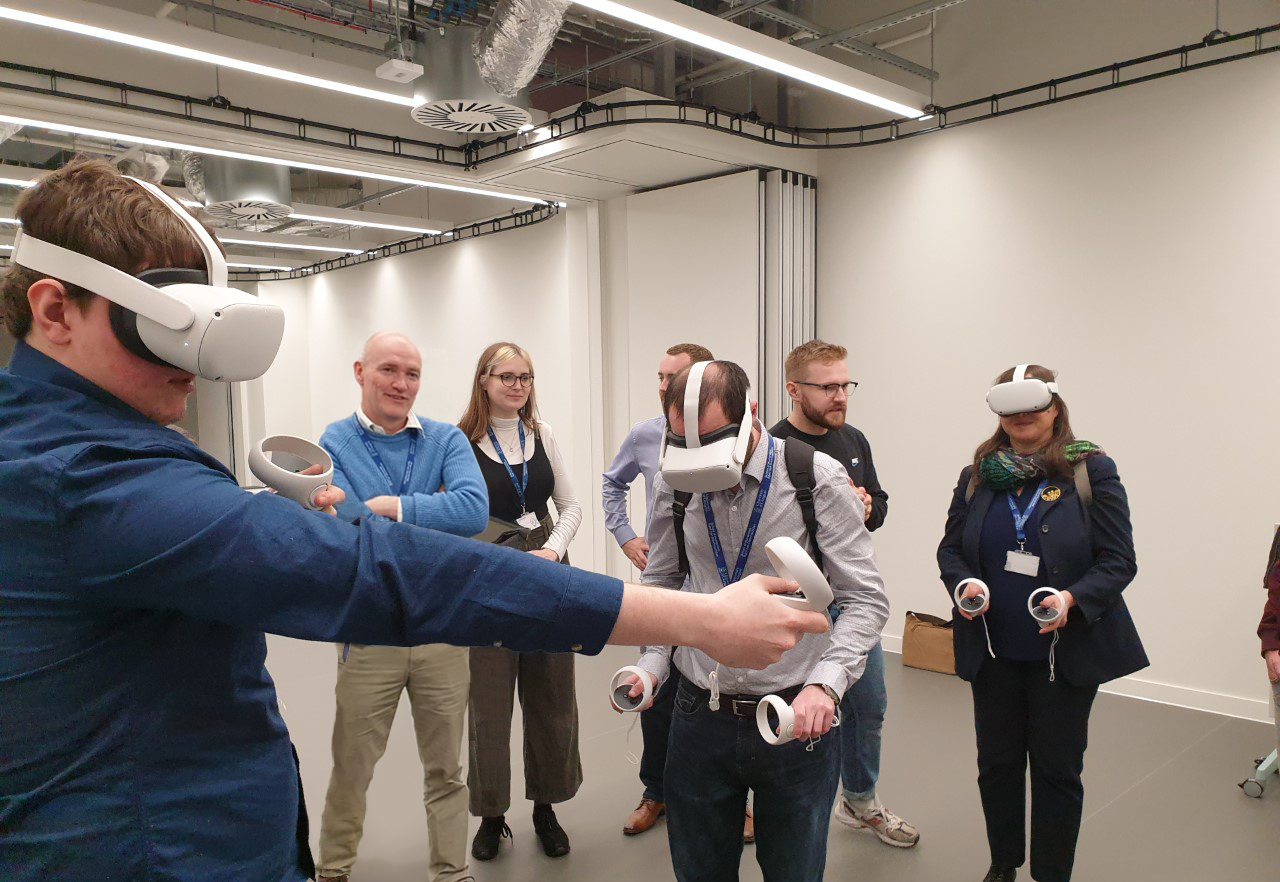 A group of people in a large room, so of them wearing VR headsets and stretching their arms out, holding small control devices