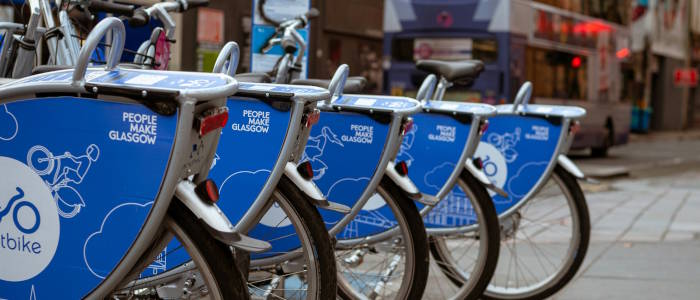 Blue Glasgow cycle hire bikes lined up at a docking station on an urban street, with city buildings and a bus in the background, representing active and sustainable travel in the city.