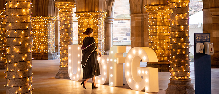 Girl in graduation gown walks through cloisters towards UofG letters