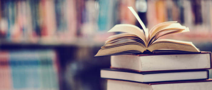 A stack of books with an open book on the top, with shelves of books in the background