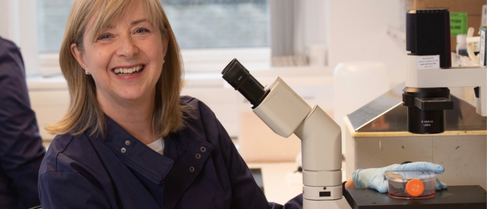 Sheila Graham wearing blue lab coat and sitting in front of a microscope and wearing latex gloves.